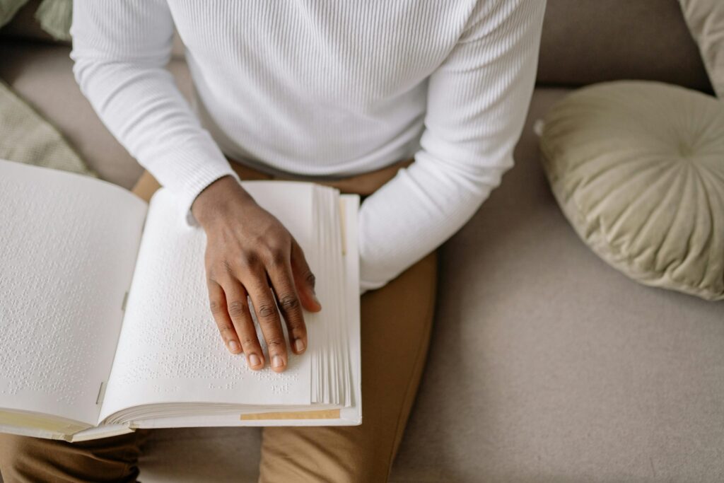 A black hand is shown reading a Braille book, with the fingers delicately gliding over the raised dots. The person is wearing a crisp white shirt, providing a clean contrast to the textured Braille page beneath their hand. The scene captures the real-life moment of someone using their sense of touch to engage with text, highlighting accessibility and the empowerment of tactile learning.